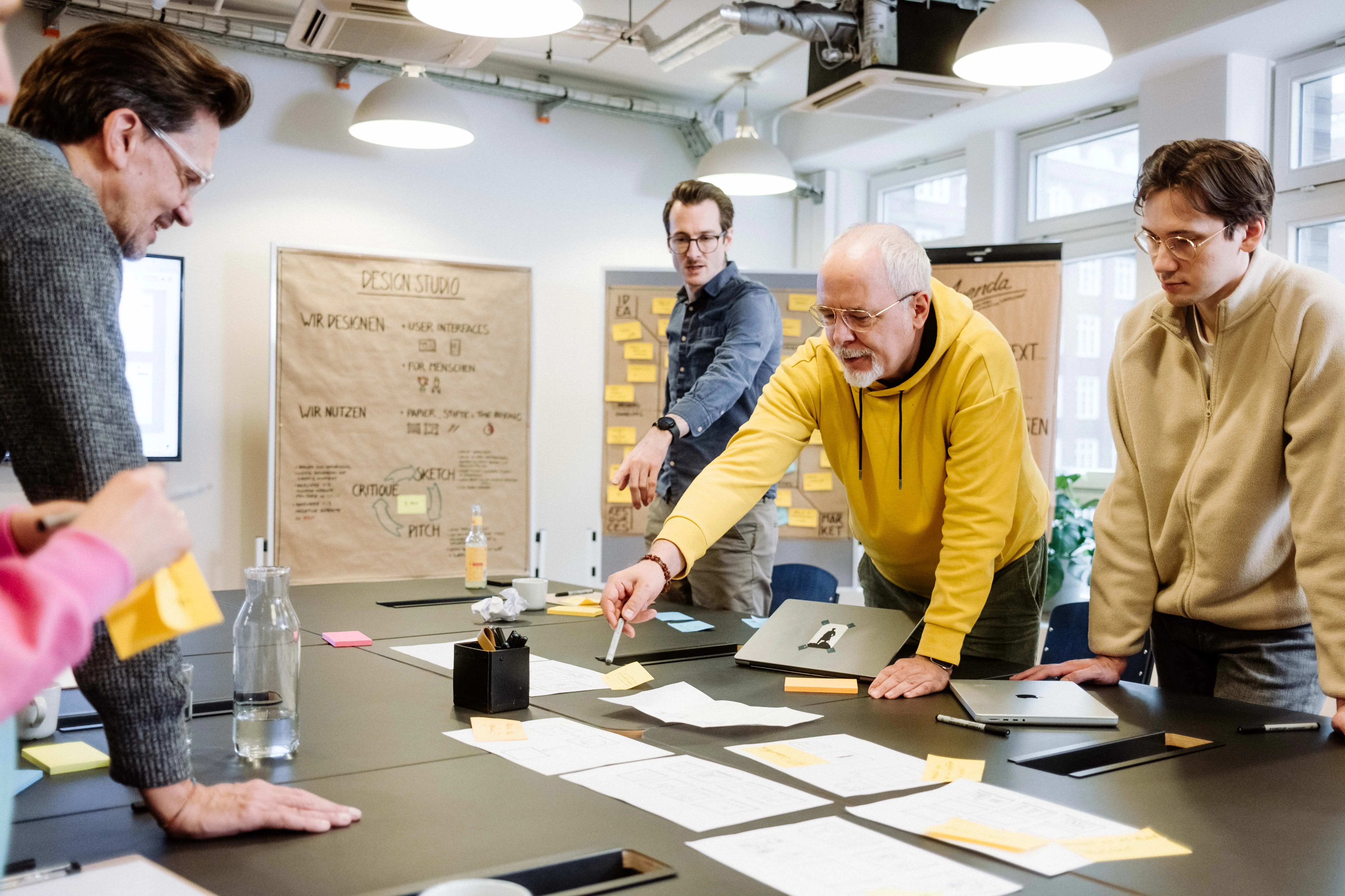 Mehrere Personen arbeiten an einem Tisch mit Skizzen, Notizzetteln und Laptops in einem Workshop.